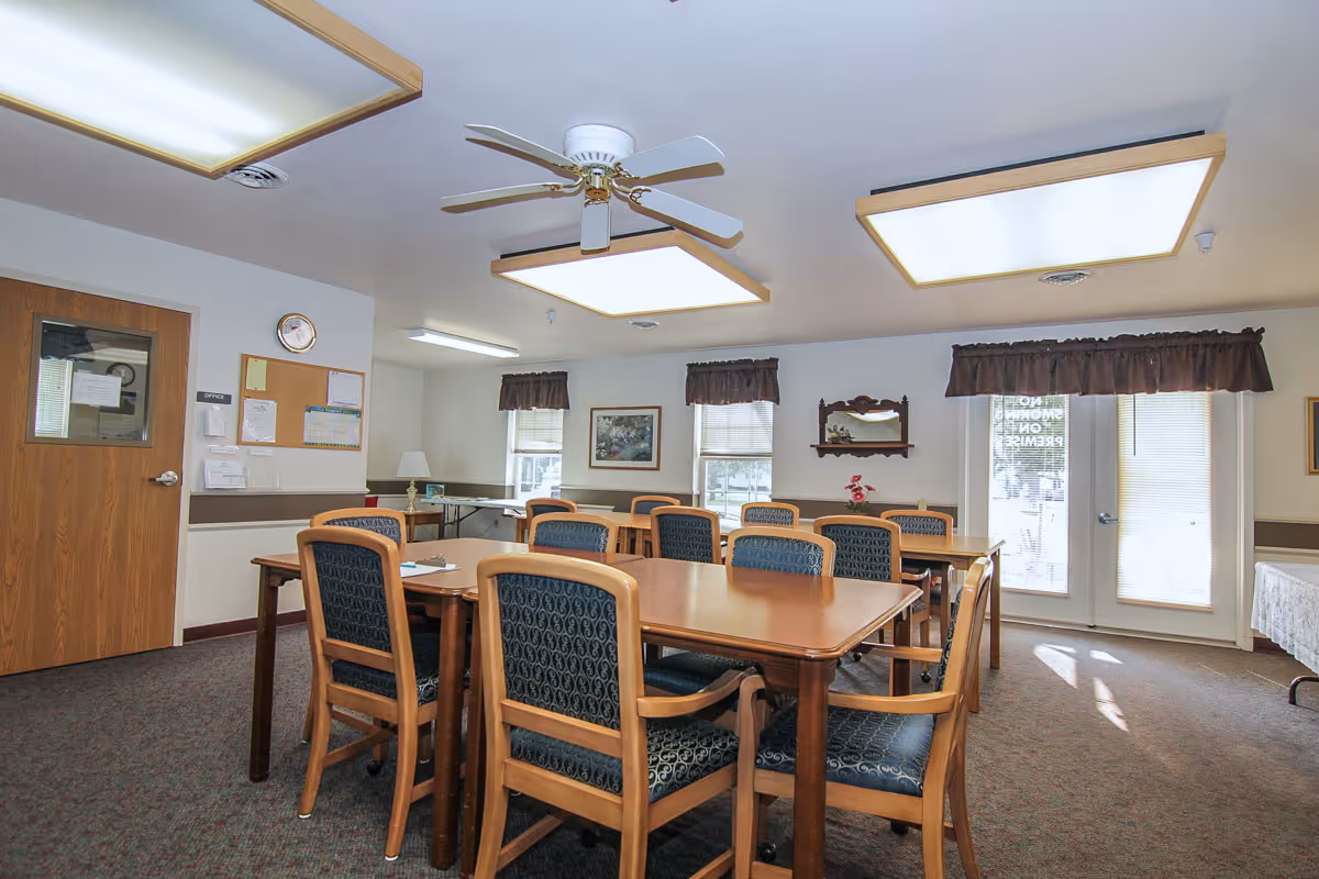 A bright communal dining room with wooden tables and upholstered chairs, ceiling fans, and large overhead lights.