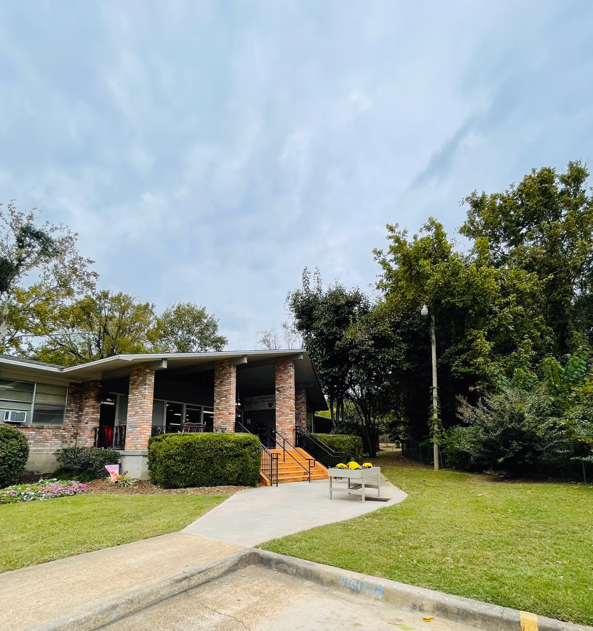 Exterior view of Natchez Rehabilitation and Healthcare Center showing a brick building with a covered entrance supported by brick columns. There are steps leading up to the entrance, surrounded by green bushes and a well-maintained lawn with trees in the background under a cloudy sky.