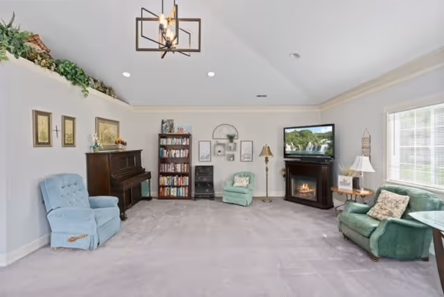 A spacious living room with light gray carpet and white walls. The room features a blue armchair, a dark wooden piano, a bookshelf filled with books, a green armchair, a floor lamp, and a corner fireplace with a TV mounted above it. There are framed pictures and decorative items on the walls and shelves, and a large window letting in natural light on the right side.