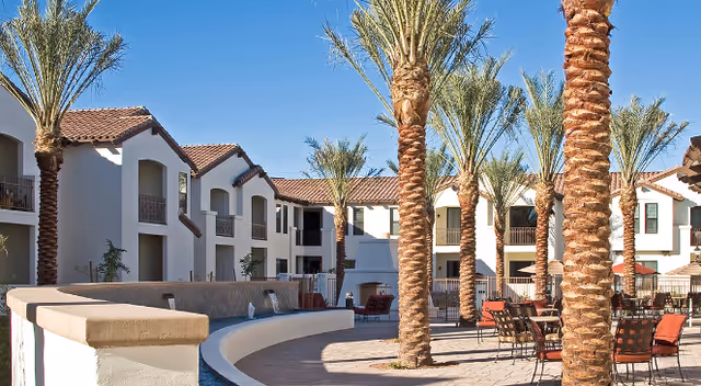 Courtyard with palm trees, a curved fountain, patio seating, and Mediterranean-style apartment buildings under a clear blue sky.