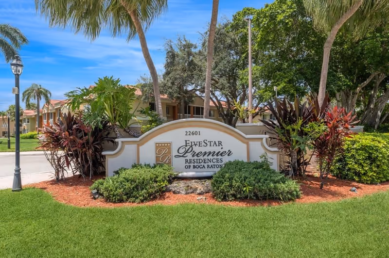Outdoor view of the entrance sign for Five Star Premier Residences of Boca Raton surrounded by green grass, shrubs, palm trees, and other plants under a blue sky.