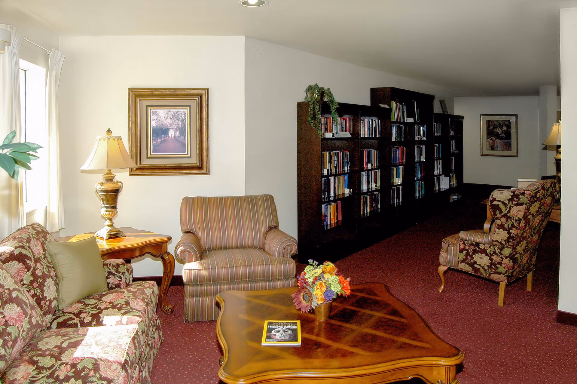 A cozy living room area with floral and striped upholstered chairs and sofa, a wooden coffee table with a flower arrangement and a book, a side table with a decorative lamp, framed artwork on the walls, and a large bookshelf filled with books along the back wall.