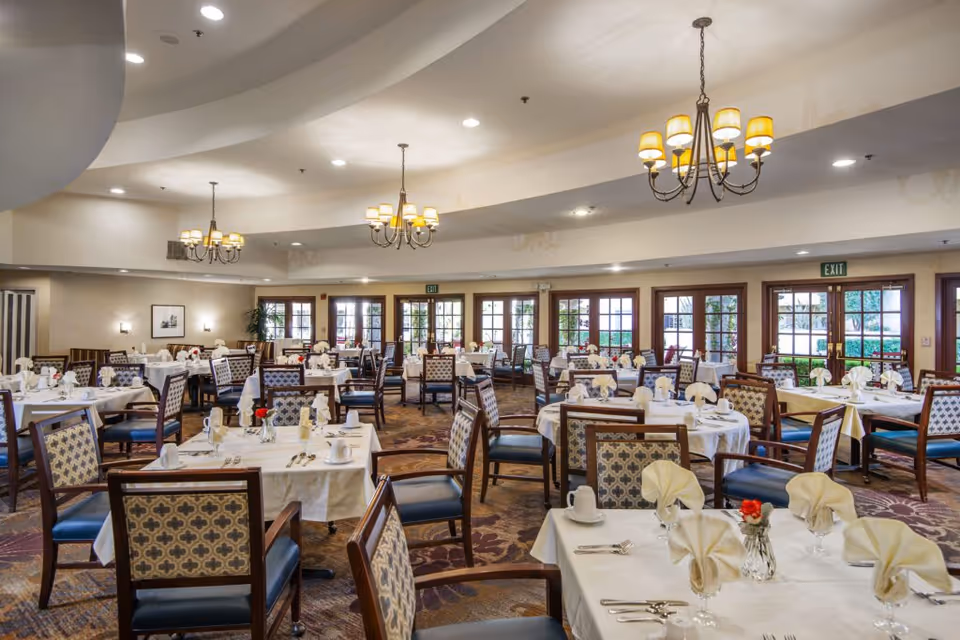 A spacious dining room with multiple tables covered in white tablecloths, each set with folded napkins, glassware, and silverware. The room features patterned chairs, chandeliers hanging from a curved ceiling, and large windows and doors letting in natural light.