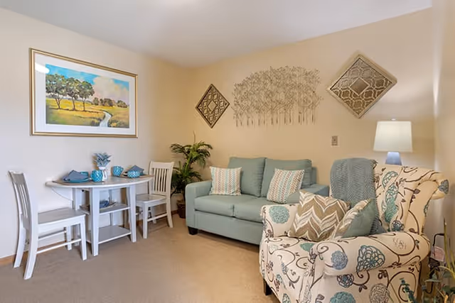 A cozy living room area featuring a light blue sofa with patterned cushions, a floral armchair with multiple pillows, a small round dining table with two chairs, decorative wall art including a framed landscape painting and metal tree sculpture, and a floor lamp providing soft lighting.