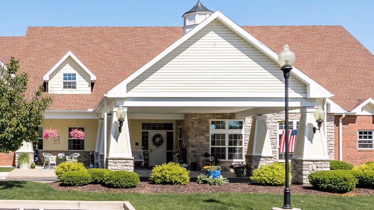 Front exterior view of a senior living facility building with a covered entrance, stone and brick facade, manicured bushes, hanging flower baskets, and an American flag displayed near the window.