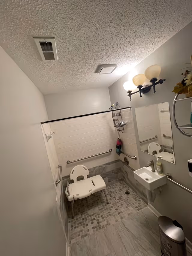 A bathroom designed for accessibility with a white shower chair, grab bars on the walls, a small sink with a soap dispenser, a mirror, and a light fixture with three bulbs. The floor has a mix of tile patterns, and there is a metal trash can in the corner.