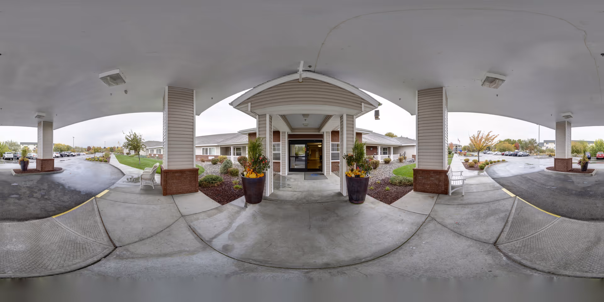 Covered entrance/porte-cochere of a senior living facility with planters, benches, and a circular driveway leading to glass double doors.