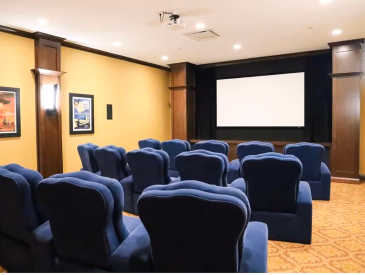 A small screening room with rows of blue upholstered chairs facing a blank projector screen and framed posters on yellow walls.