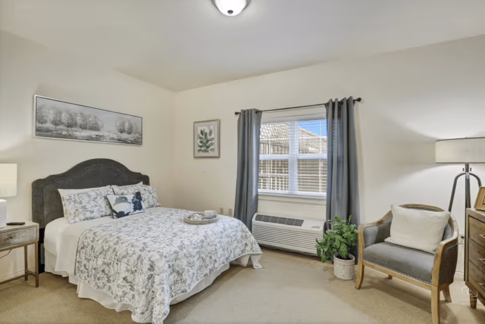 A cozy bedroom with a bed featuring a floral patterned comforter and pillows, a wooden nightstand with a lamp, a window with gray curtains, a gray armchair with a white cushion, a floor lamp, a wooden dresser, and framed artwork on the walls.