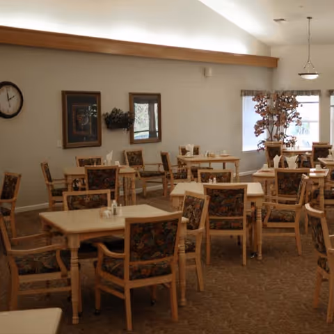 A dining room with multiple wooden tables and chairs featuring floral upholstery. The room has beige walls, a clock on the wall, framed artwork, and a large window with a potted plant nearby. The lighting is soft with a hanging light fixture visible.
