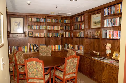 A cozy library room with wooden bookshelves filled with books lining the walls. In the center, there is a round wooden table surrounded by six upholstered chairs with floral patterned fabric. The room also features framed artwork on the walls and decorative items on the shelves and countertop.