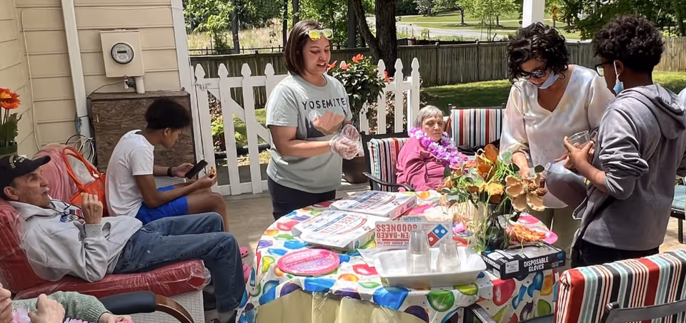 A group of people gathered outdoors around a table covered with a colorful tablecloth. There are pizza boxes, disposable gloves, and cups on the table. Some individuals are seated on cushioned chairs, while others are standing and interacting. The setting appears to be a patio area with a white picket fence and greenery in the background.