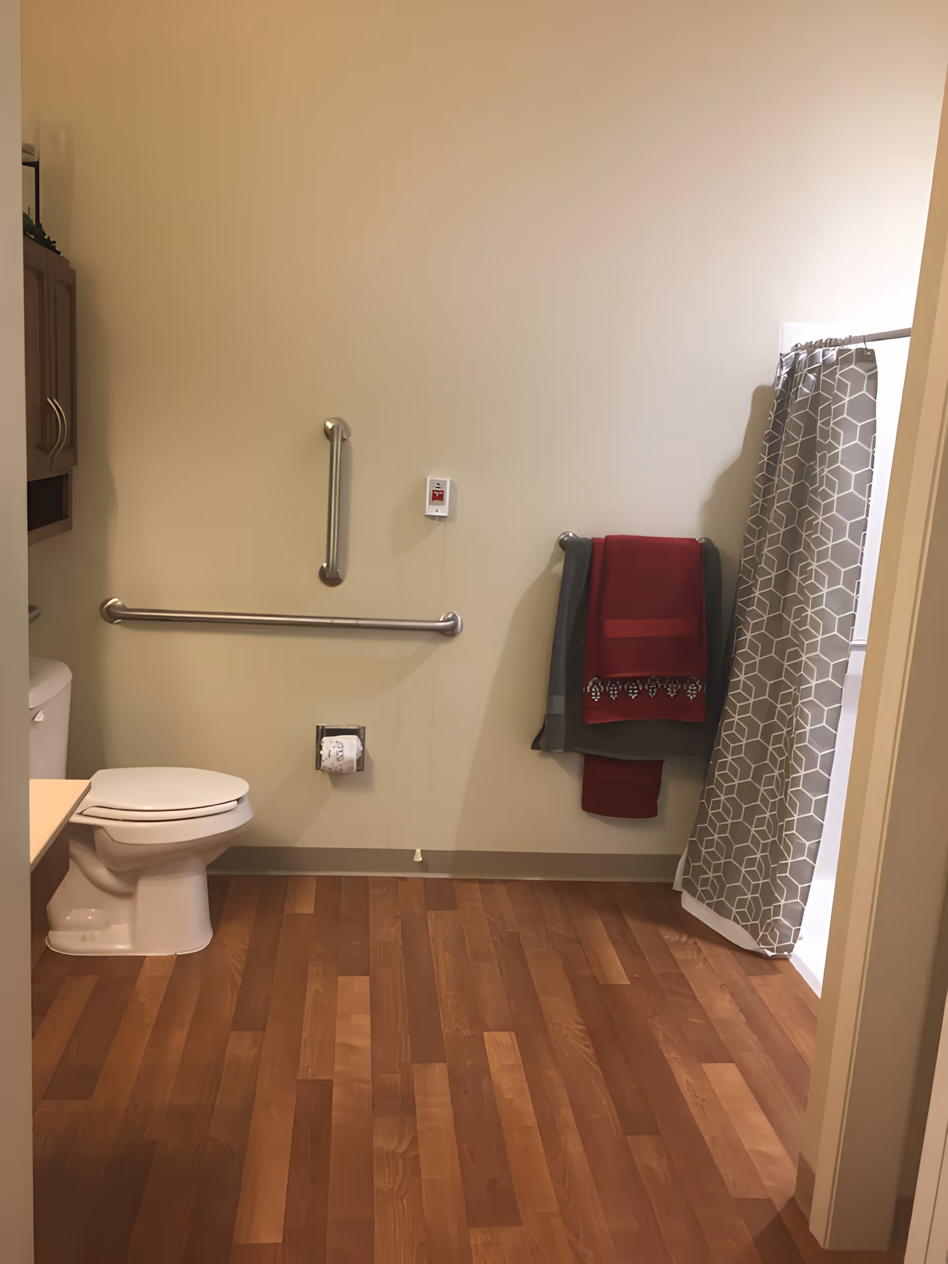 A bathroom with wooden flooring, a white toilet, grab bars on the wall, a towel rack with red and gray towels, and a shower area with a gray and white patterned curtain.