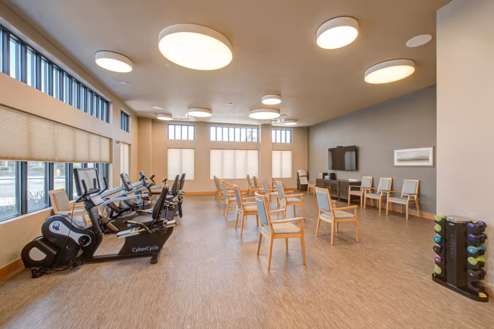 A spacious exercise room with several stationary exercise bikes lined up near large windows on the left. Multiple wooden chairs with light blue cushions are arranged in rows facing a wall-mounted TV and a cabinet. The room has large round ceiling lights, light-colored walls, and a rack of colorful dumbbells on the right side.