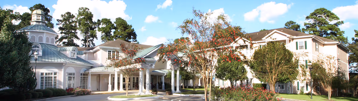 Exterior view of King's Preserve at Kingwood, a large senior living facility with white walls, green shutters, and a distinctive cupola on the roof. The building is surrounded by trees, shrubs, and a well-maintained garden under a blue sky with scattered clouds.