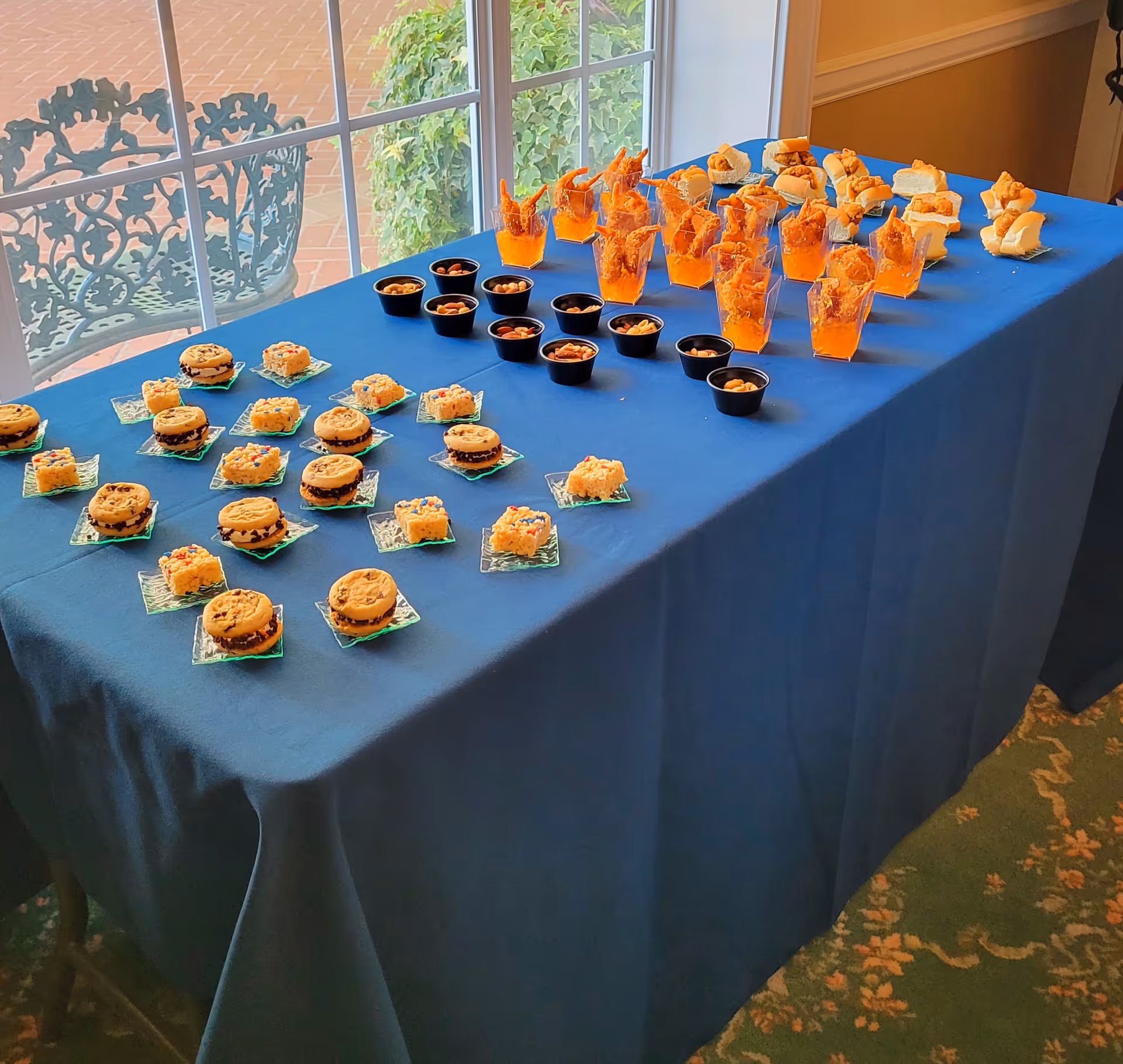 A table covered with a blue tablecloth displaying an assortment of snacks including mini sandwiches, small cups with nuts, shrimp in orange sauce, and various cookies and treats. The table is positioned near a window with a view of outdoor patio furniture and greenery.