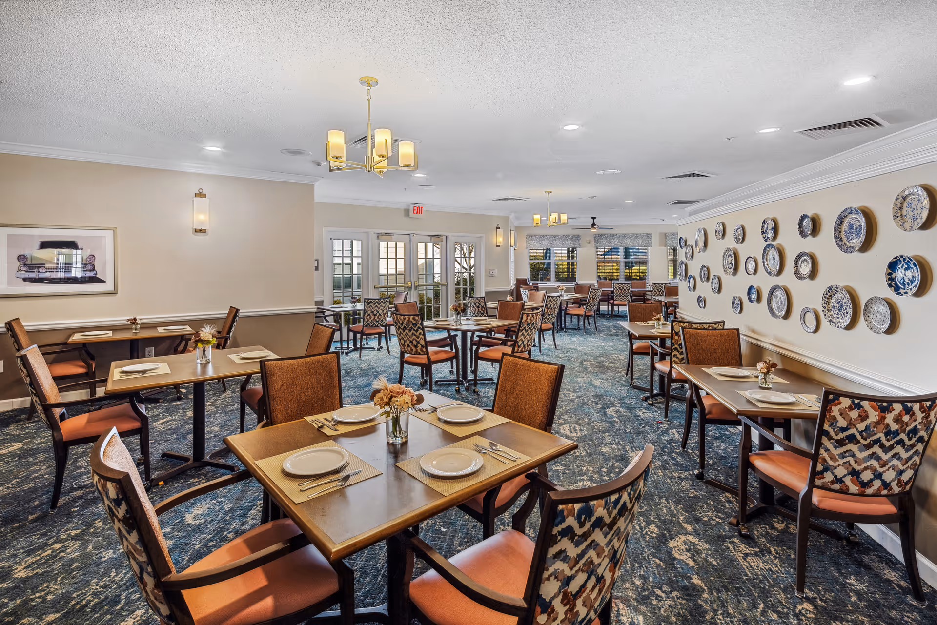 A well-lit dining room with multiple tables set with plates, utensils, and small flower vases. The room features patterned chairs, a blue and beige carpet, and decorative plates mounted on one wall. Large windows and glass doors allow natural light to enter the space.