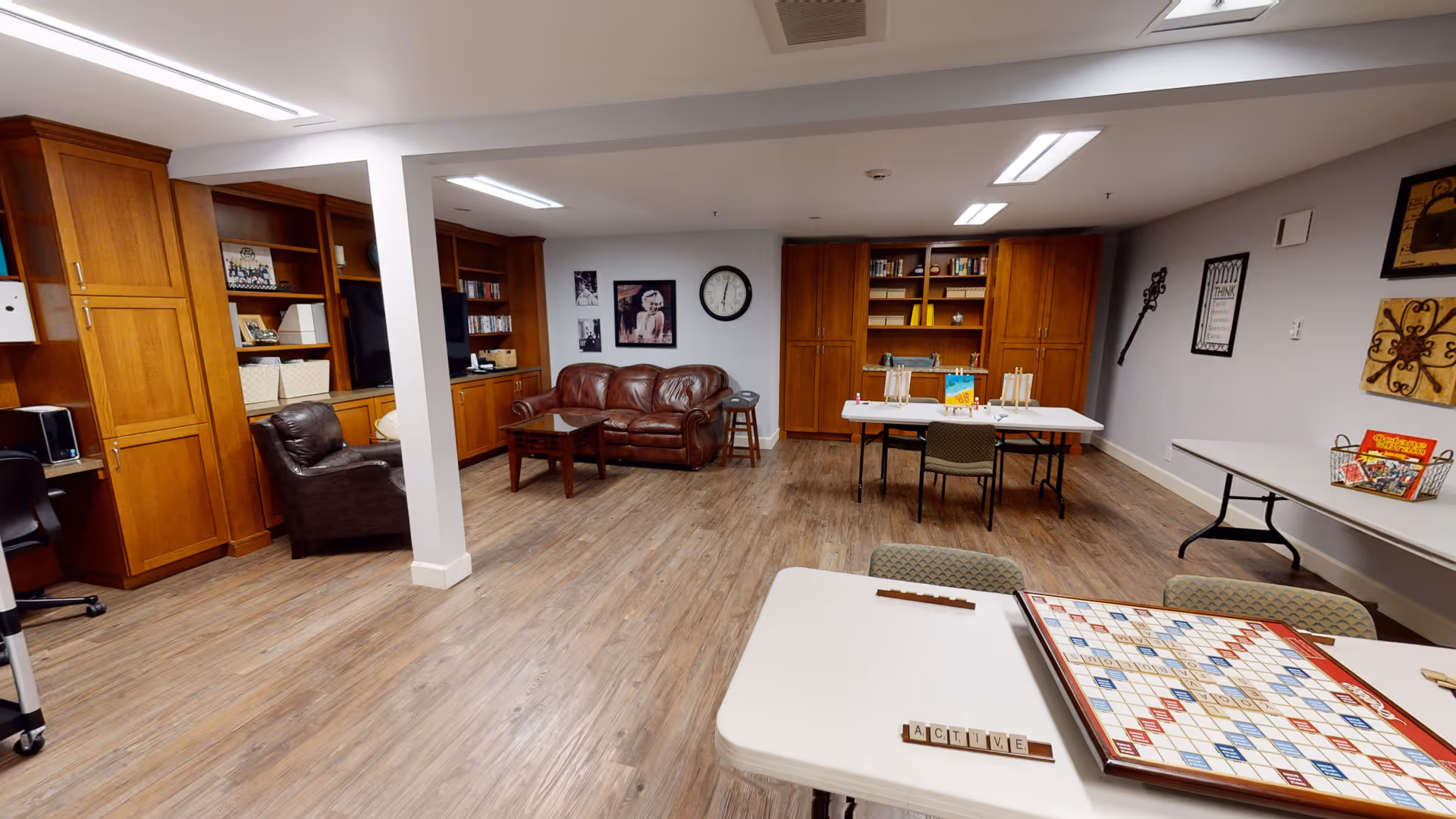A spacious common room with wooden flooring and built-in wooden cabinets along the walls. The room features a brown leather couch, a matching armchair, several tables with chairs, and a Scrabble board game set up on one table. The walls are decorated with framed pictures and a clock.