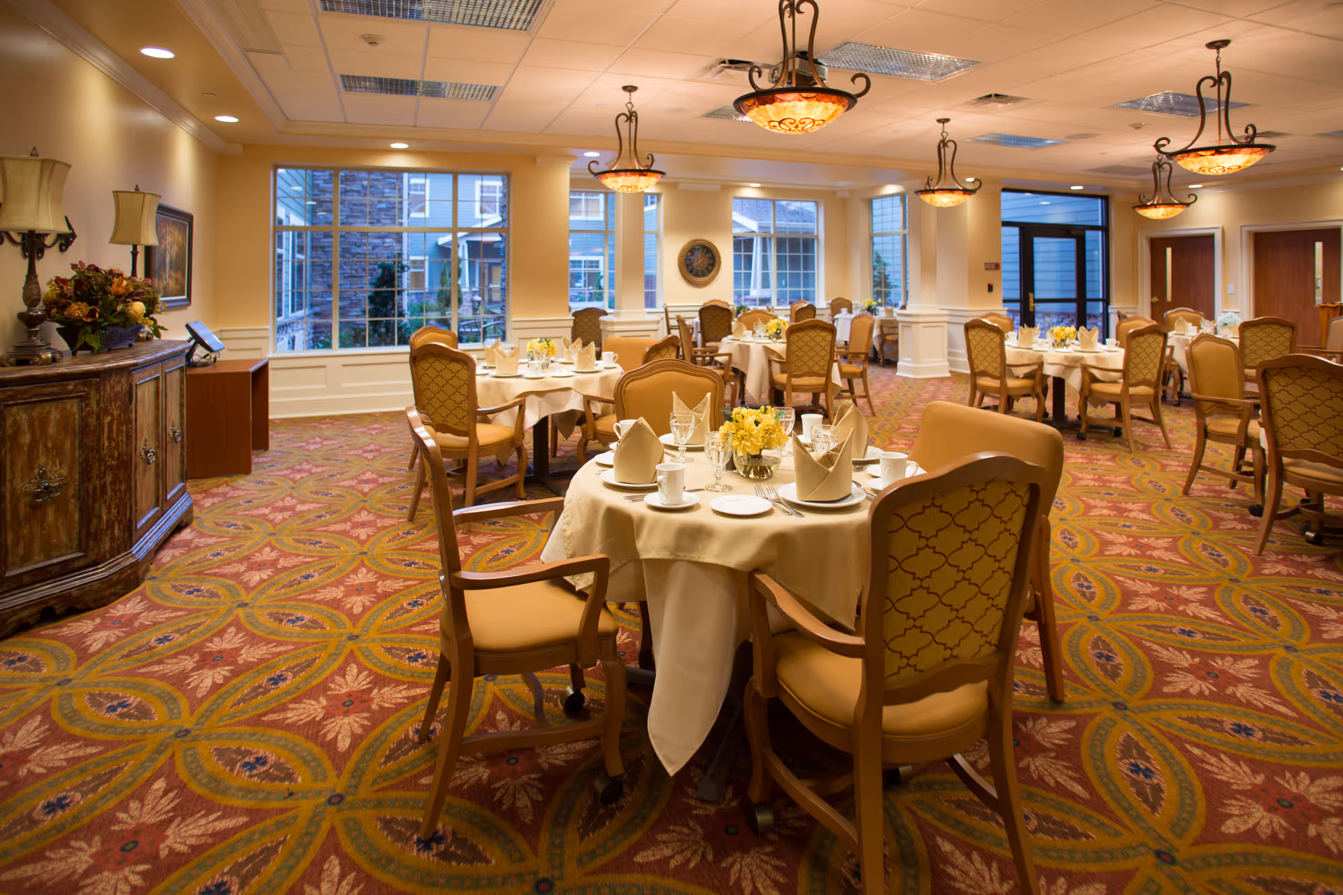 A well-appointed dining room with round tables set for meals, upholstered chairs and decorative ceiling lights.
