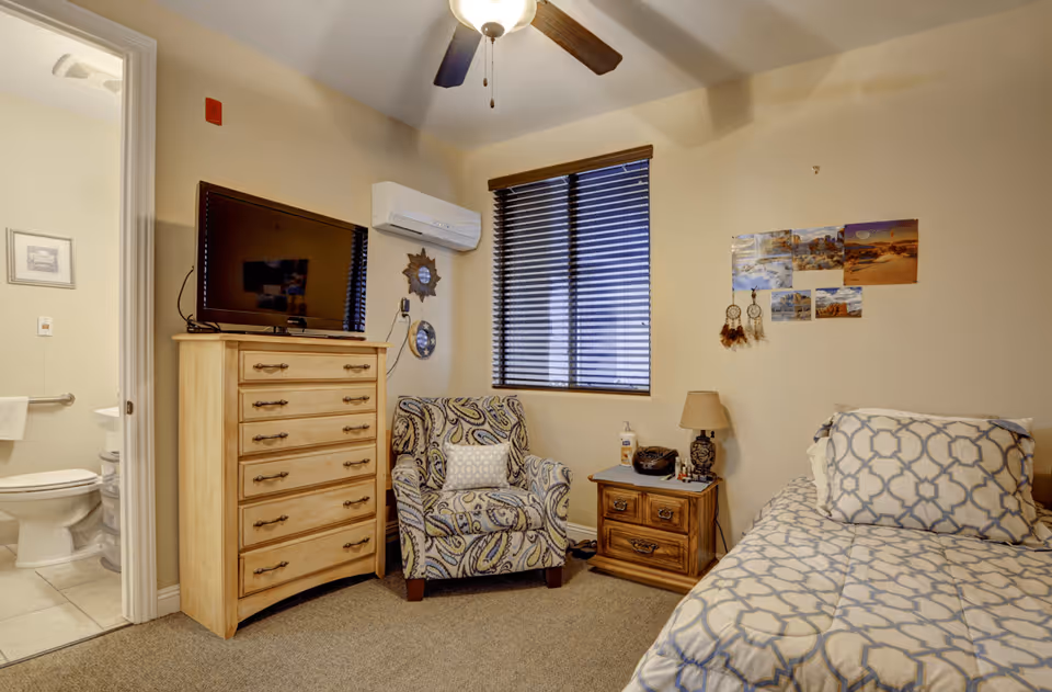 A cozy bedroom with a patterned armchair, a wooden dresser with a flat-screen TV on top, a nightstand with a lamp and personal items, and a bed with patterned bedding. There is a window with closed blinds and several pictures and dreamcatchers on the wall. A bathroom is visible through an open door.