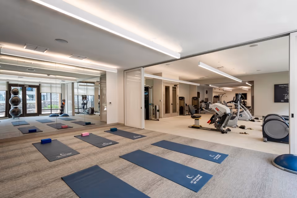 Bright fitness/yoga studio with multiple mats and blocks on the floor, a mirrored wall, and exercise equipment visible in the adjoining room.