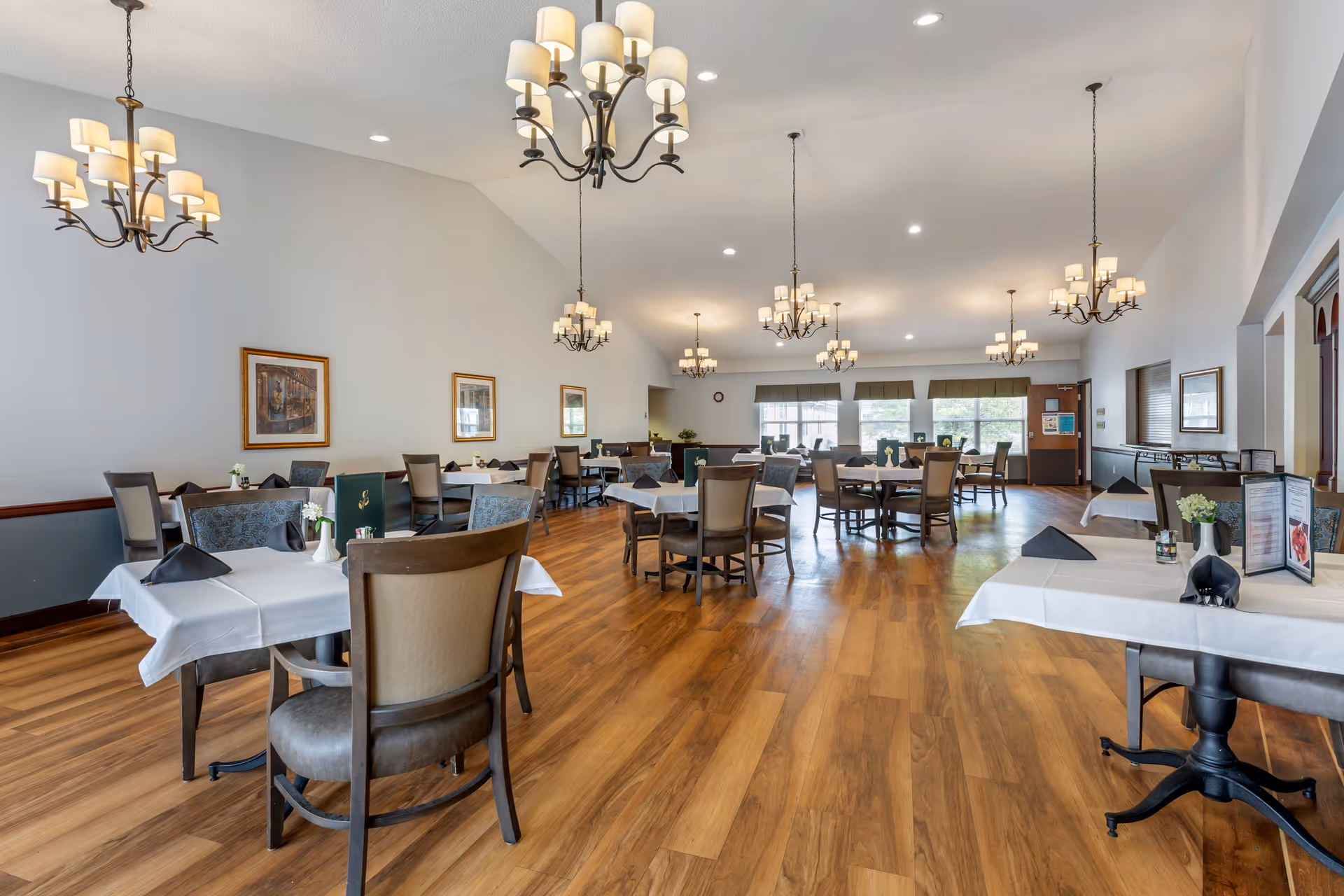 Spacious dining room with multiple tables covered in white tablecloths, each set with chairs and small decorative items. The room features wooden flooring, several chandeliers hanging from the ceiling, framed artwork on the walls, and large windows letting in natural light.
