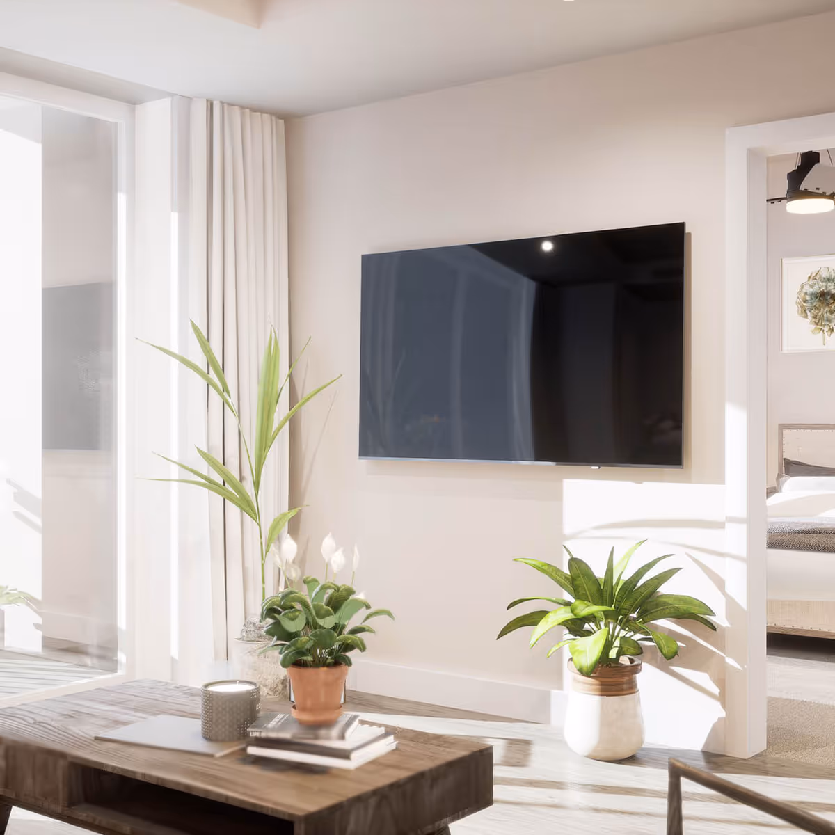 Bright living room with a wall-mounted flat-screen TV, a wooden coffee table and several potted plants near a doorway to a bedroom.
