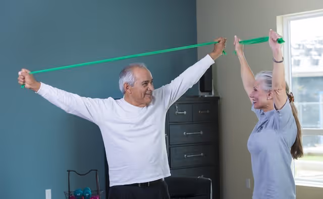 An elderly man and woman exercising indoors using a green resistance band, both smiling and stretching the band above their heads in a room with a window and a black cabinet in the background.