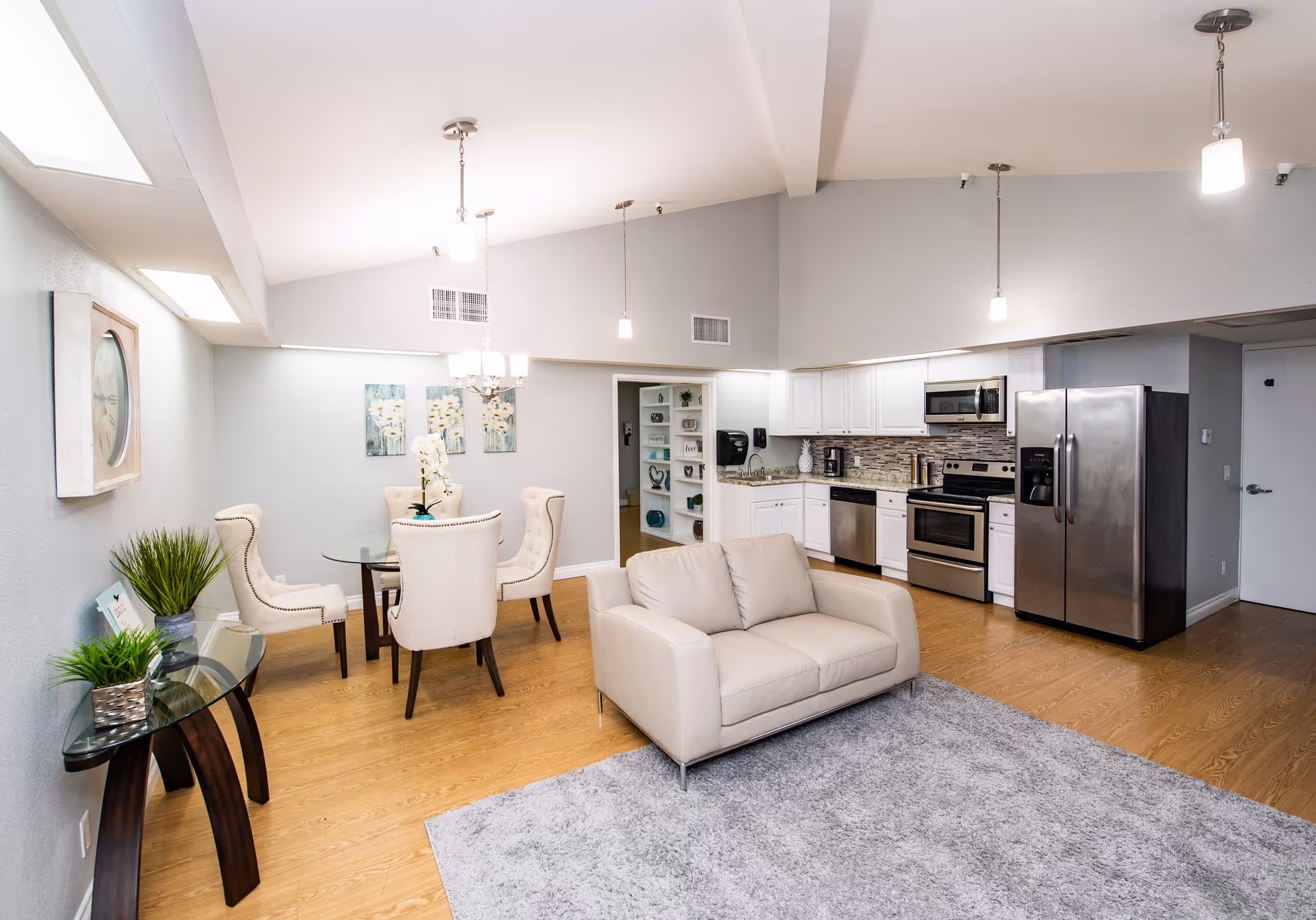 A bright and modern open-concept living space featuring a small beige loveseat on a gray rug, a glass dining table with four upholstered chairs, and a kitchen area with stainless steel appliances including a refrigerator, oven, microwave, and dishwasher. The room has light wood flooring, white walls, and ceiling lights hanging above the kitchen and dining area. Decorative plants and wall art add a cozy touch.