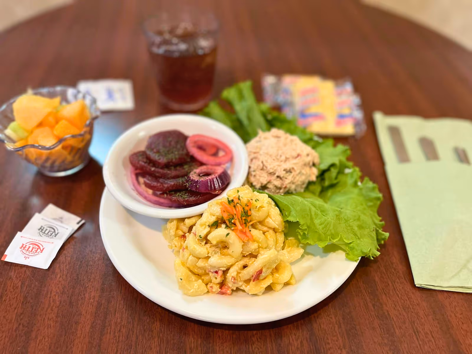 A meal served on a white plate with macaroni salad, a scoop of tuna salad on lettuce, and a small bowl of sliced beets and onions. Next to the plate is a small bowl of mixed fruit, a glass of iced tea, packets of salt and pepper, and a folded green napkin on a wooden table.