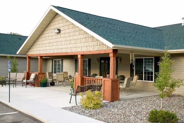 Exterior view of a single-story building with a green roof and beige siding. The building has a covered porch with wooden posts and outdoor seating including chairs and benches. There are small trees and shrubs planted in a rock garden near the sidewalk and a handicapped parking sign visible in the parking area.