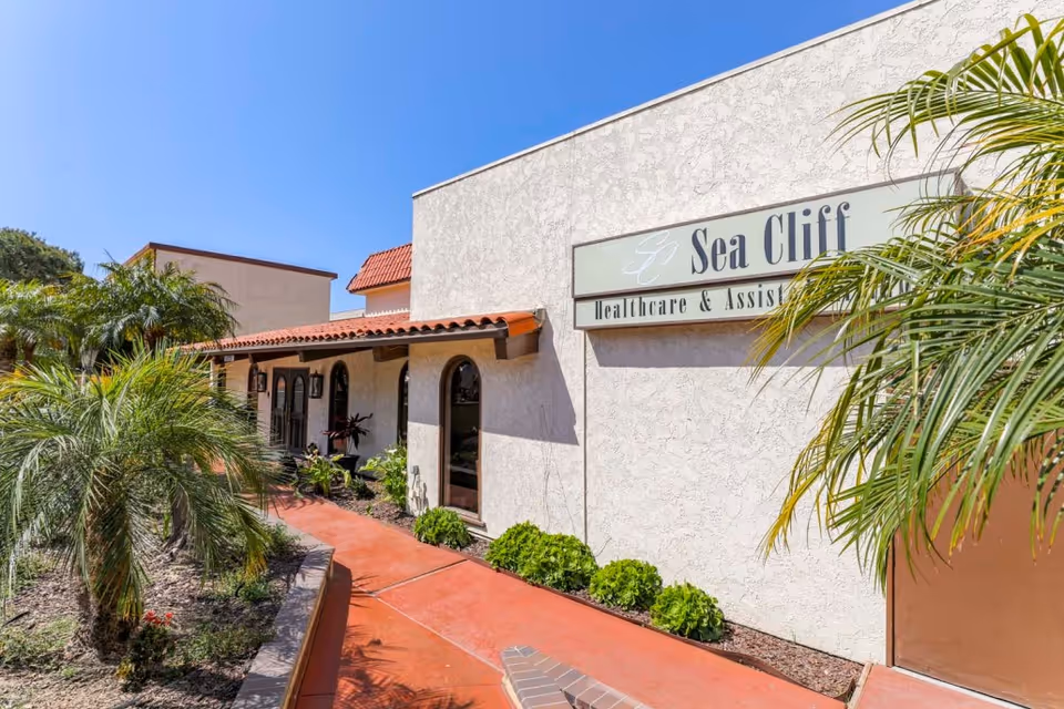 Exterior view of Sea Cliff Assisted Living facility with a beige stucco wall, a sign displaying the facility name, a red tiled roof over the entrance, and landscaped greenery including palm trees and bushes along a red concrete walkway under a clear blue sky.