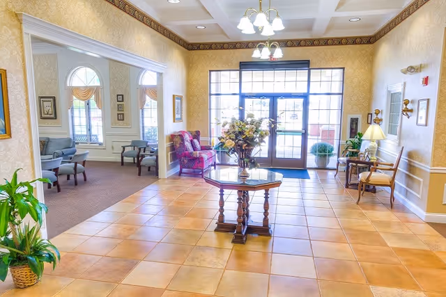 Bright senior living lobby with a tiled floor, a central table topped with flowers, seating areas, and glass entrance doors.