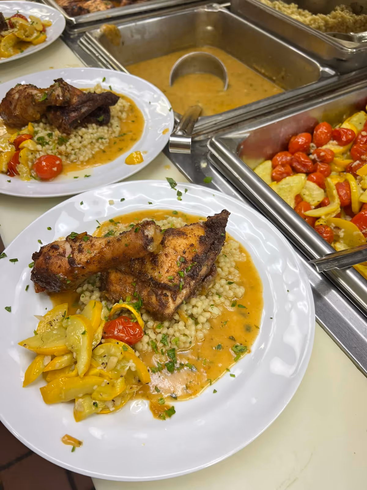 Two plates of food with grilled chicken drumsticks and thighs served over a bed of couscous or small pasta pearls with a light sauce, accompanied by sautéed yellow squash and cherry tomatoes. In the background, there are stainless steel food trays with more cooked vegetables and sauce.