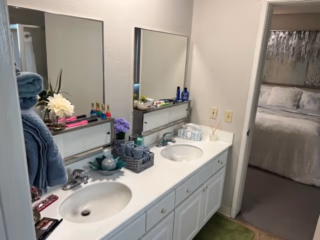 A bathroom vanity with two sinks, each with a mirror above. The countertop has various items including towels, flowers, toiletries, and decorative pieces. To the right, an open doorway leads to a bedroom with a bed covered in light-colored bedding and a large piece of artwork above the headboard.