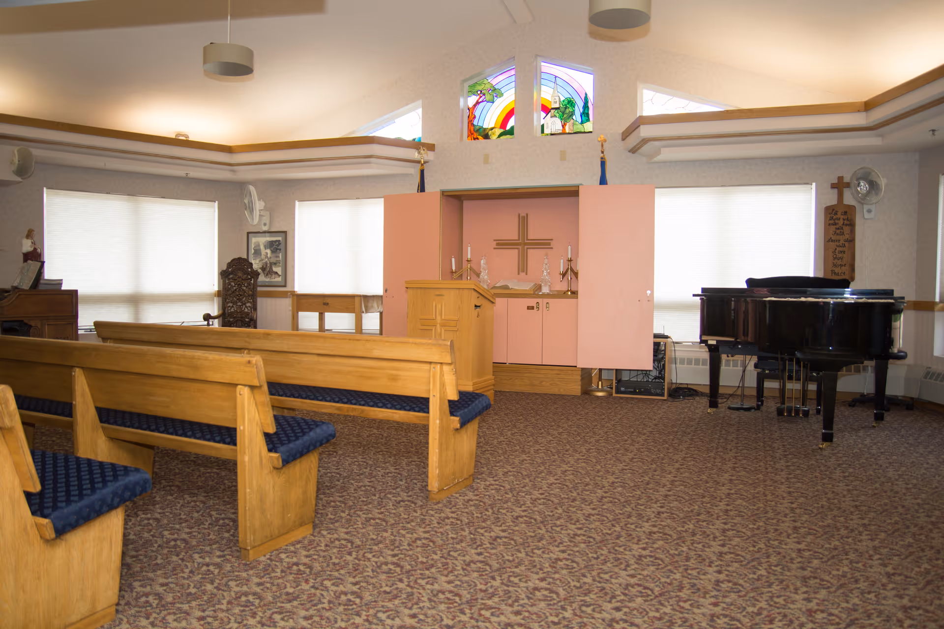 Chapel-style interior with wooden pews, a lectern/altar area and a grand piano beneath stained glass windows.