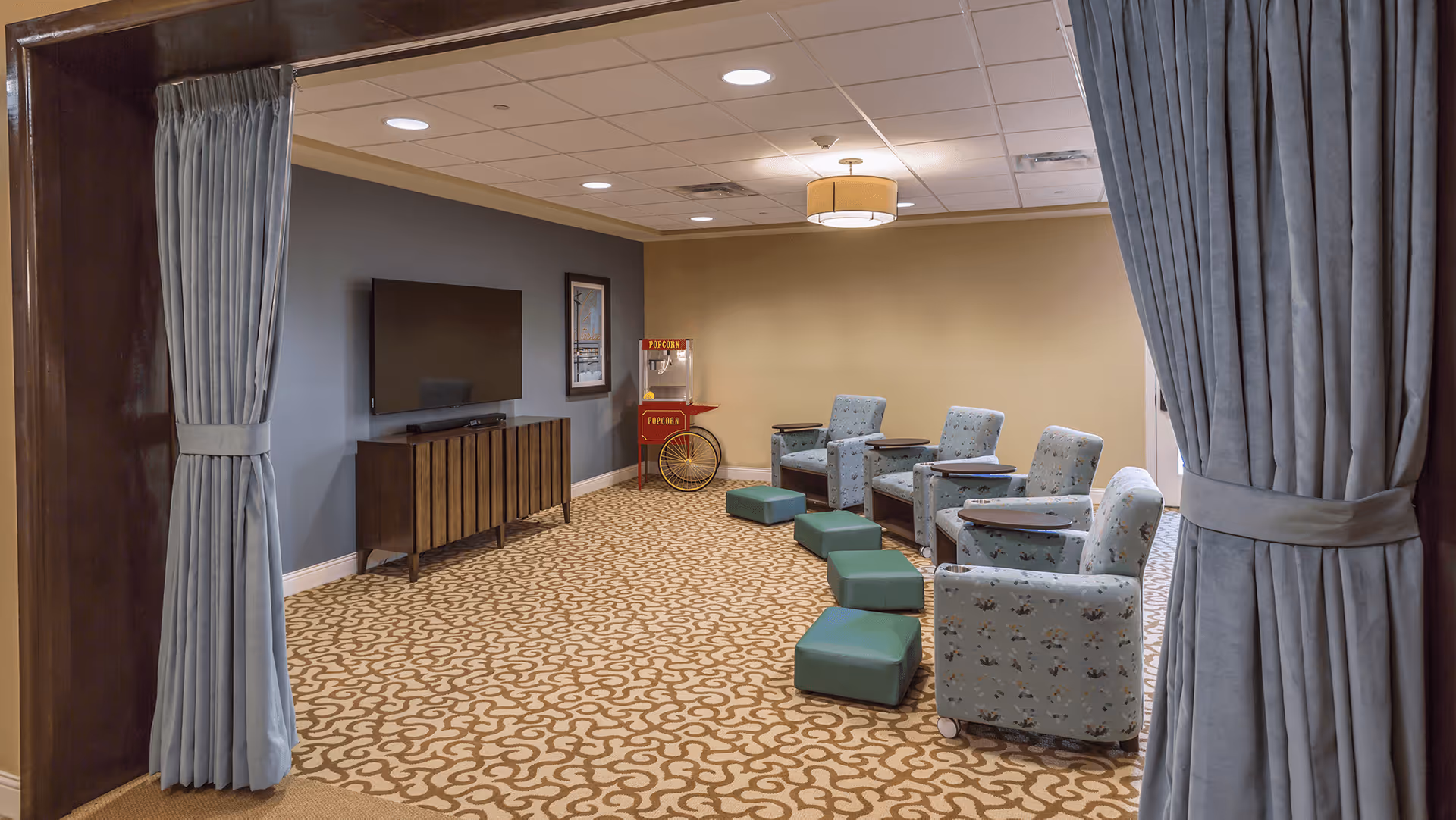 A cozy lounge area with four patterned armchairs each paired with a small green ottoman. The room has a patterned carpet, a flat-screen TV mounted on a blue accent wall above a wooden console, and a vintage-style popcorn machine in the corner. The space is framed by light blue curtains tied back on either side.