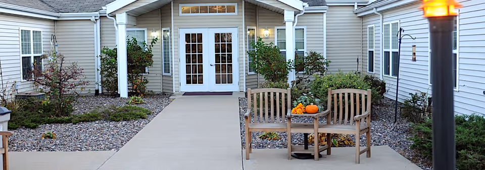 Outdoor seating area at the entrance of a senior living facility with two wooden chairs and a small table holding a pumpkin and flowers, surrounded by landscaping with bushes and rocks, and a building with beige siding and white-framed windows and doors in the background.