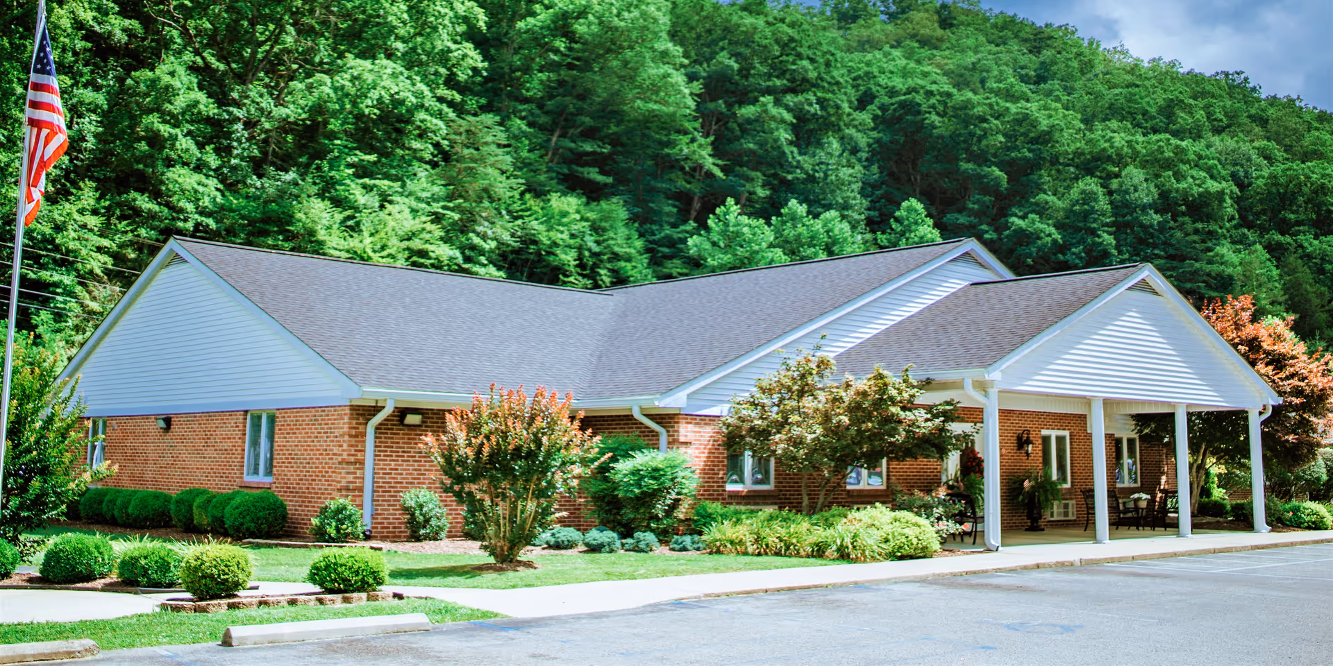 Single-story brick senior living facility with a covered entrance, landscaped shrubs and lawn, and an American flag in front.
