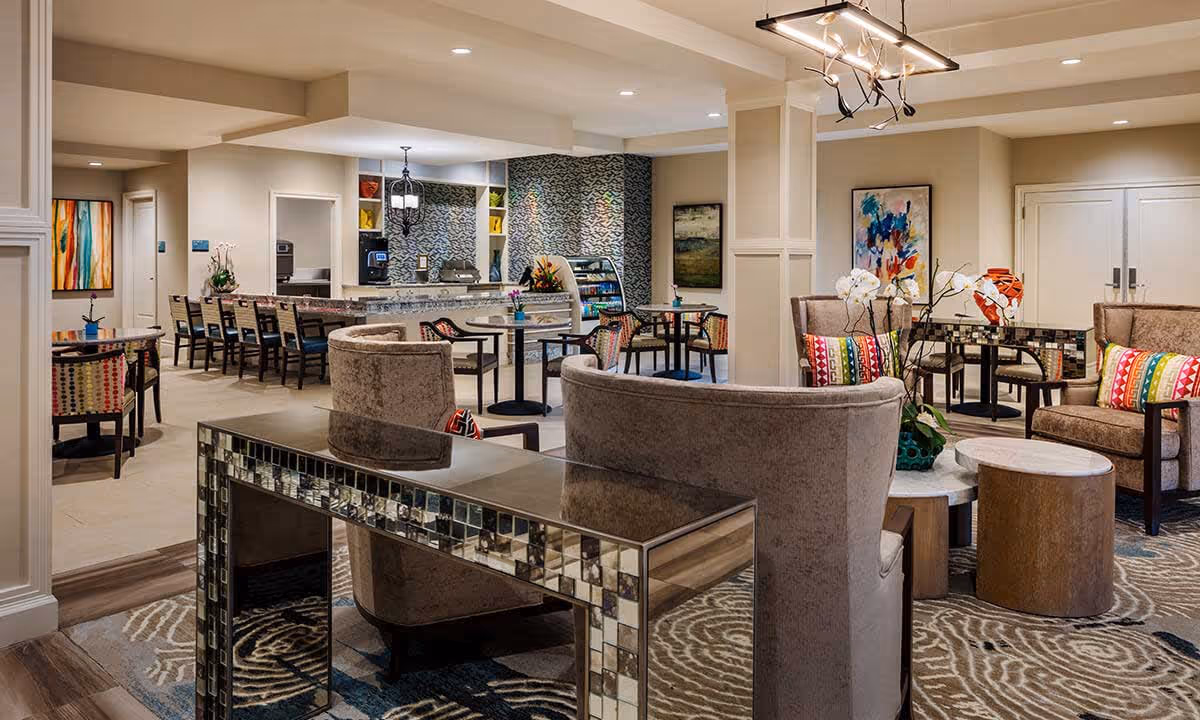 Spacious, brightly lit senior living lounge with upholstered seating, small tables, colorful pillows, and a café-style counter and dining area in the background.