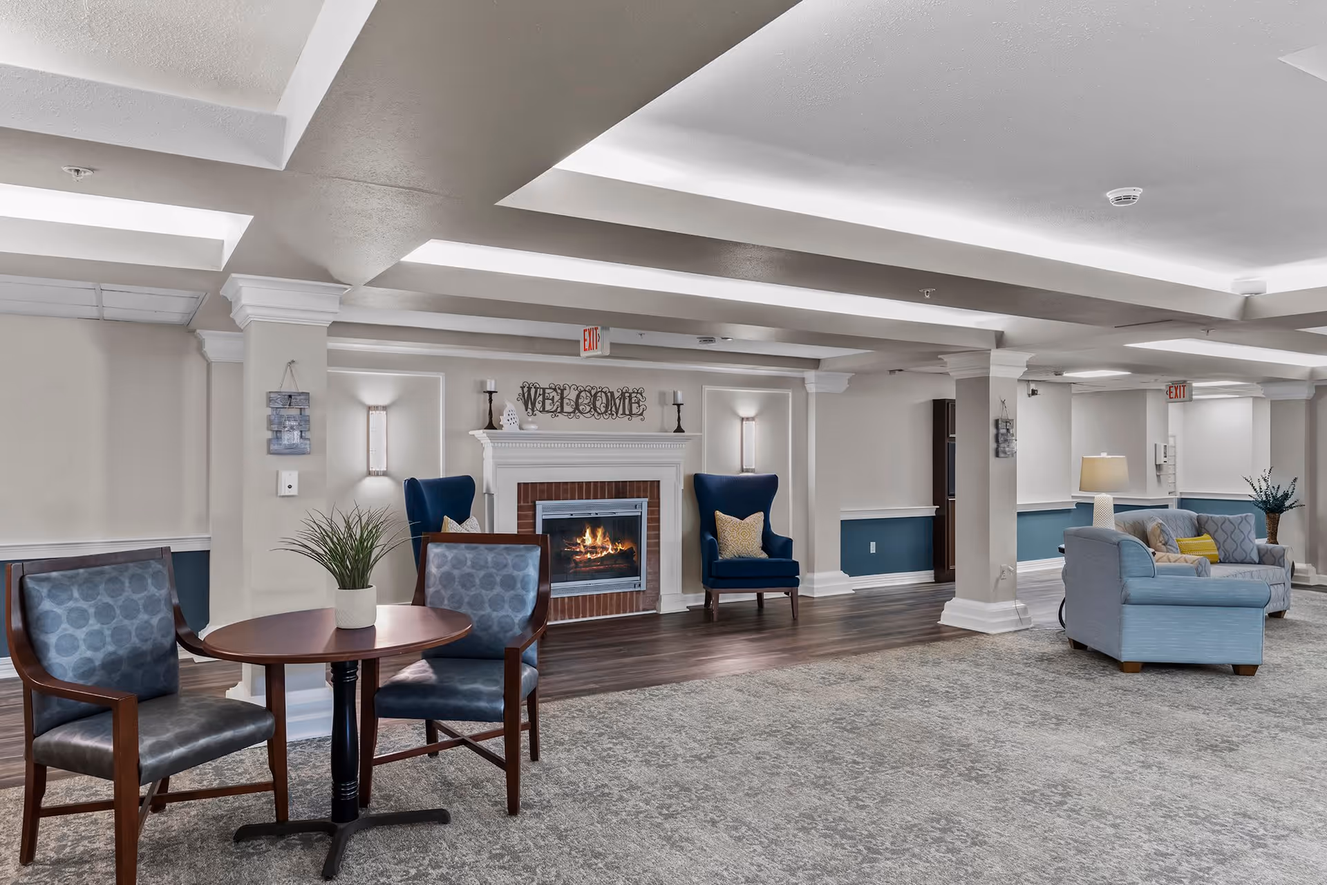 A spacious and well-lit living room area in a senior living facility featuring a fireplace with a 'WELCOME' sign above it, two blue armchairs with yellow pillows on either side of the fireplace, a round wooden table with two blue cushioned chairs, and a light blue sofa with a yellow pillow. The room has a combination of carpet and wood flooring, white walls with blue wainscoting, and ceiling beams with recessed lighting.