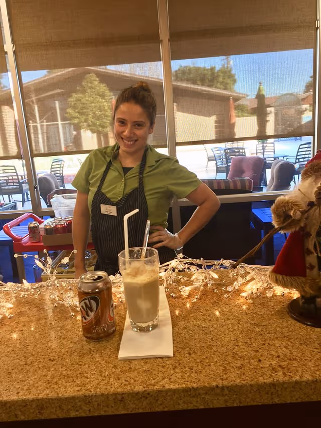 A smiling woman wearing a green shirt and black apron stands behind a counter with a can of A&W root beer and a glass with a straw and spoon on a napkin. Behind her, there are large windows with shades partially down, showing outdoor patio furniture and a building outside.