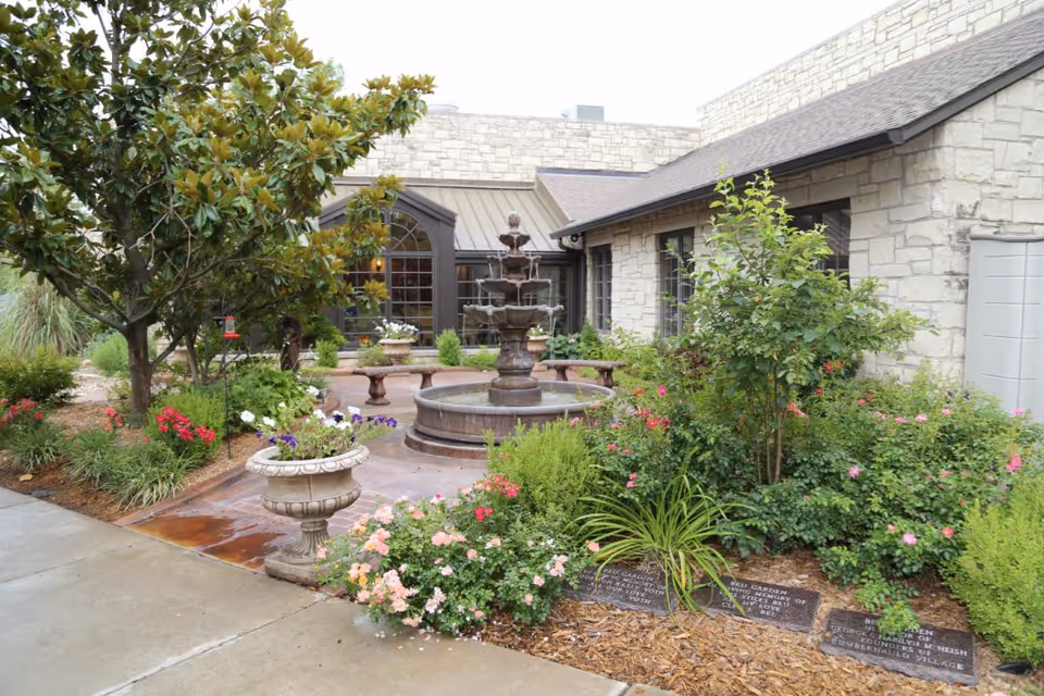 A landscaped outdoor garden area with a multi-tiered water fountain in the center, surrounded by various flowering plants and shrubs. There are stone benches around the fountain and a stone building with large windows in the background. The garden includes decorative planters and a paved walkway.