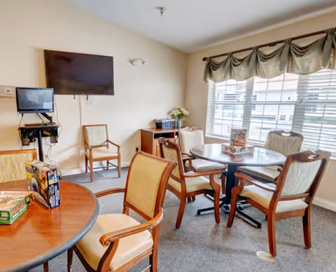 A well-lit room with two round wooden tables surrounded by cushioned chairs. On the tables, there are board games. The room has a large window with green valance curtains allowing natural light to enter. A flat-screen TV is mounted on the wall, and there is a small cabinet with a basket and flowers on top.