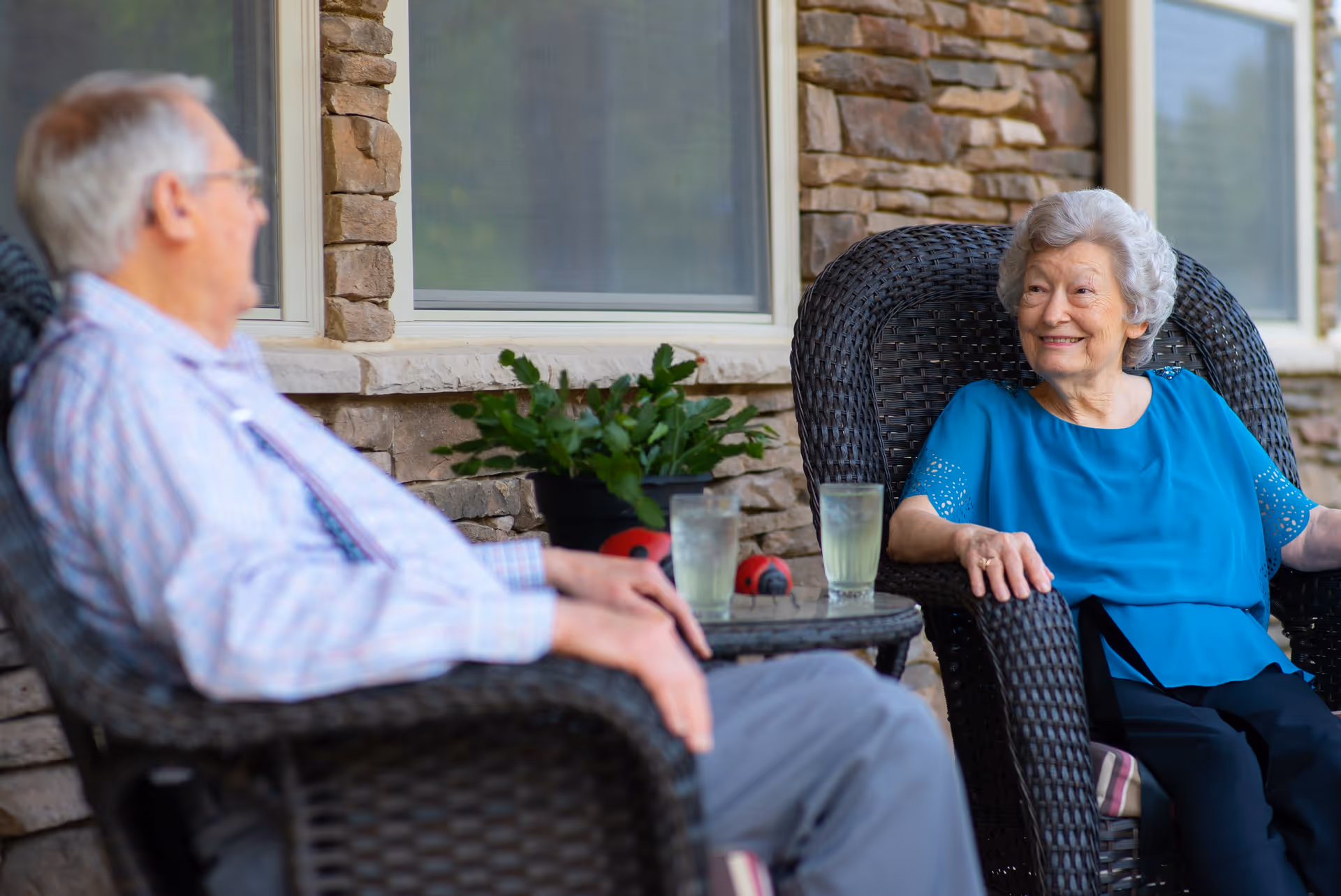 An elderly man and woman sitting in wicker chairs on a patio outside a stone building, smiling and conversing with each other. There is a small table between them with two glasses of a light-colored beverage and a potted plant.