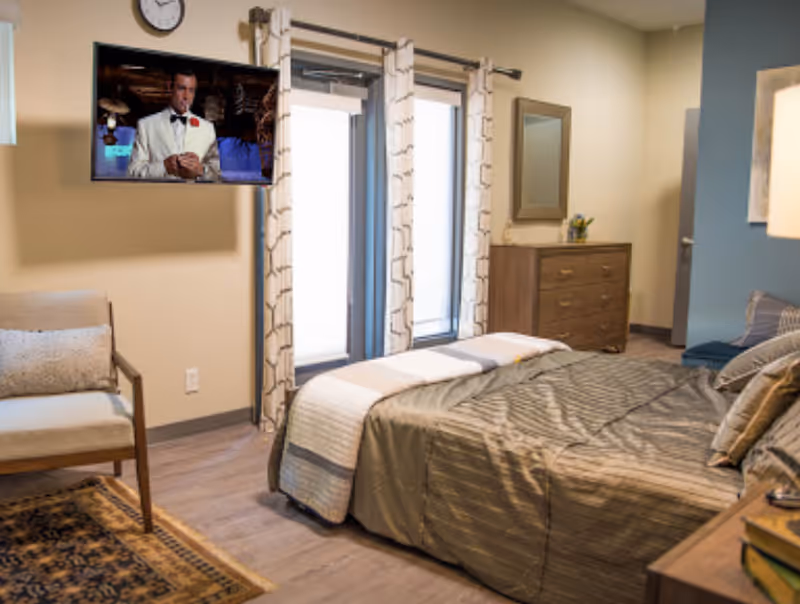 A cozy bedroom with a neatly made bed covered in a gray striped comforter and multiple pillows. There is a wooden dresser with a mirror above it, a chair with a cushion, and a wall-mounted TV showing a man in a white suit. The room has large windows with patterned curtains letting in natural light.