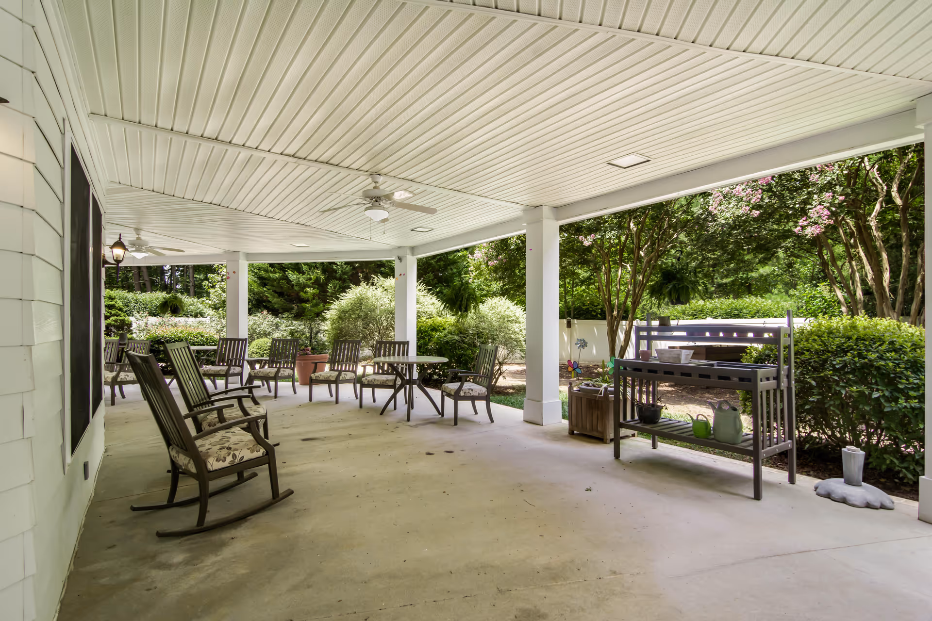 Covered outdoor patio area with several wooden rocking chairs and tables. There is a gardening shelf with pots and watering cans on the right side. The patio overlooks a garden with green bushes and trees with pink flowers.