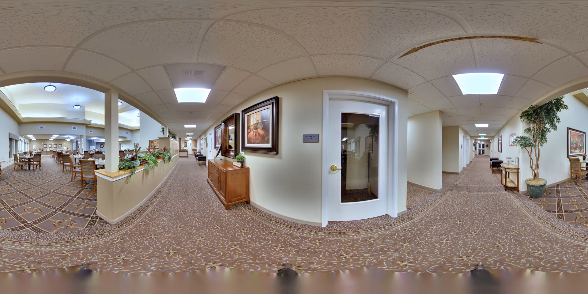 Panoramic view of a senior living facility interior hallway with patterned carpet, beige walls, and ceiling tiles. To the left is a dining area with multiple tables and chairs, separated by a half wall with plants. The hallway features framed artwork, a wooden cabinet, and a door with a sign. There are potted plants and additional seating along the hallway.