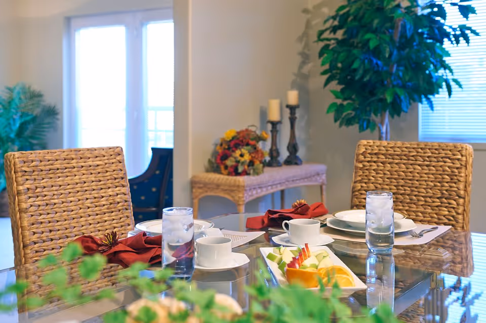 A bright dining room with a glass table set with plates, cups, glasses of water, a fruit platter and woven chairs, with plants and decorative candles in the background.