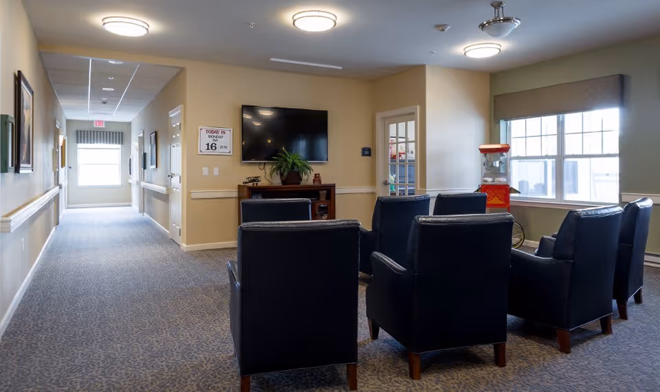 A common area in an assisted living facility with six dark blue armchairs arranged in two rows facing a wall-mounted flat screen TV. There is a wooden cabinet beneath the TV with a potted plant and decorative items. A popcorn machine is positioned near a large window with blinds. The room has beige walls, carpeted floor, and ceiling lights. A hallway extends to the left with handrails and framed pictures on the walls.